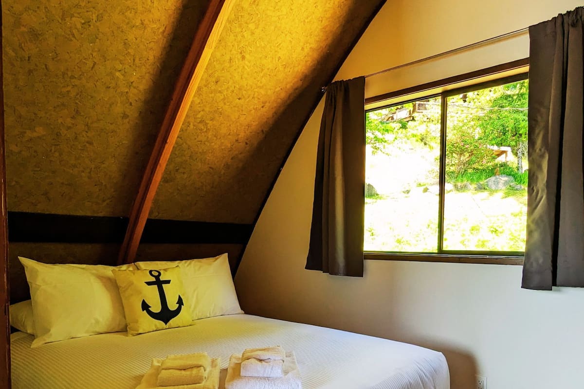 A single bed with a wooden frame, a pillow with an anchor design, and a window with curtains.