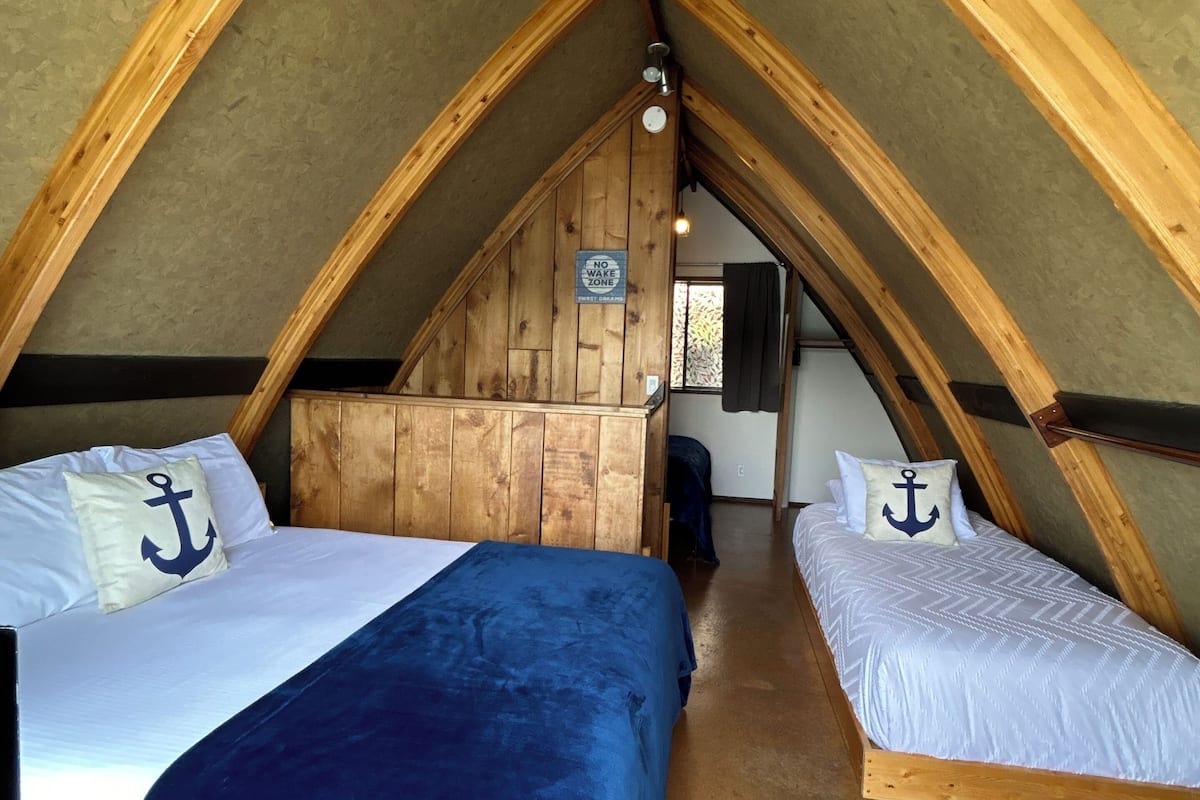 A room with a vaulted ceiling, wooden beams, two beds with navy blue and white bedding, and a sign indicating a 'no wake zone.'