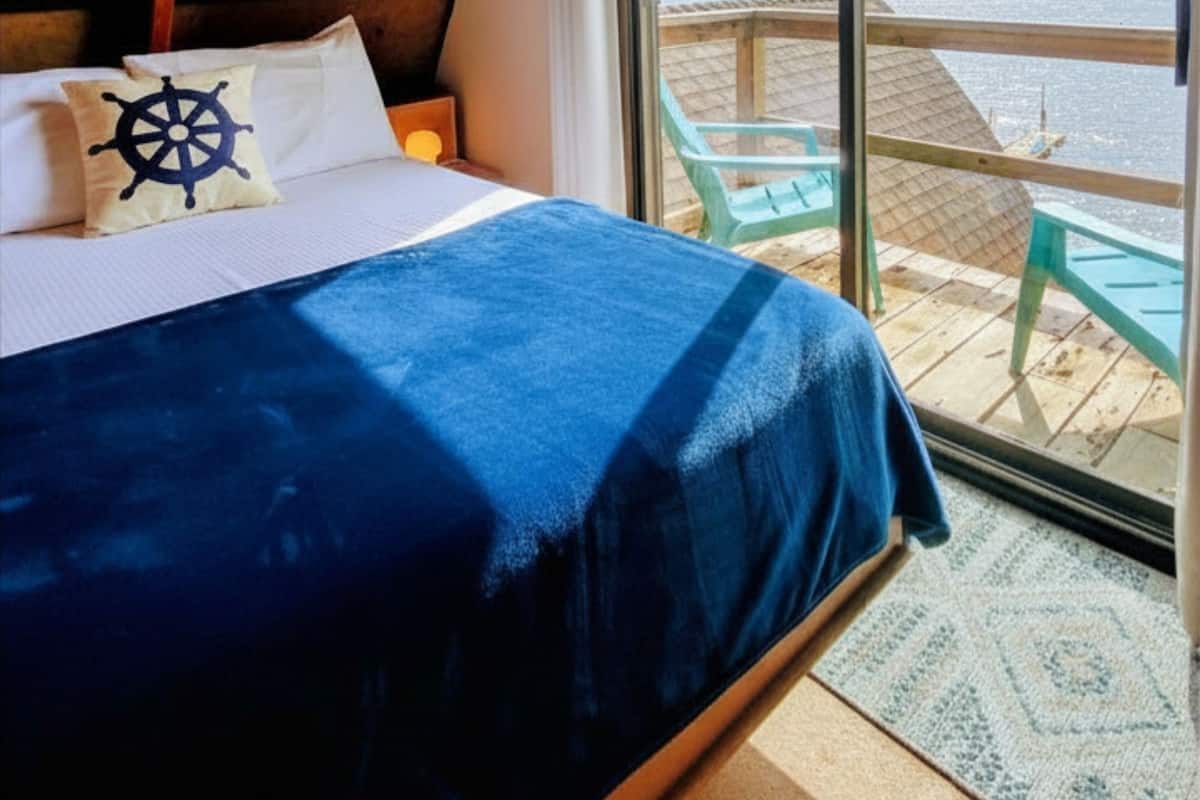 A bedroom with a bed, a view of the water, and a deck with chairs.
