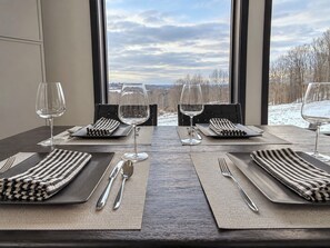A dining table set for two with a view of a snowy landscape through large windows. - Hust Haus | Sweeping Catskills Views on 20 Acres (Jeffersonville)