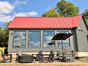 A small building with a red roof, large windows, and outdoor seating with chairs and a table. - Hust Haus | Sweeping Catskills Views on 20 Acres (Jeffersonville)