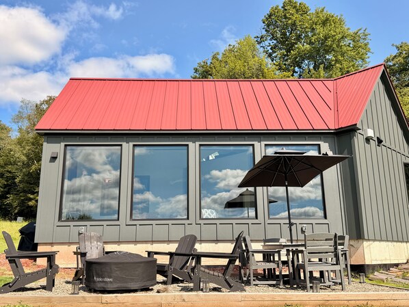 A small building with a red roof, large windows, and outdoor seating with chairs and a table. - Hust Haus | Sweeping Catskills Views on 20 Acres (Jeffersonville)