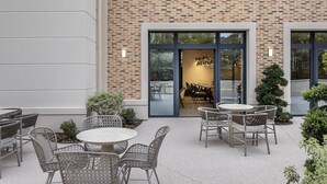 An outdoor patio area with round tables and wicker chairs, a brick wall, and a glass door leading inside. - Best Western Plus Hotel Saint-Roch Orleans (Orleans)