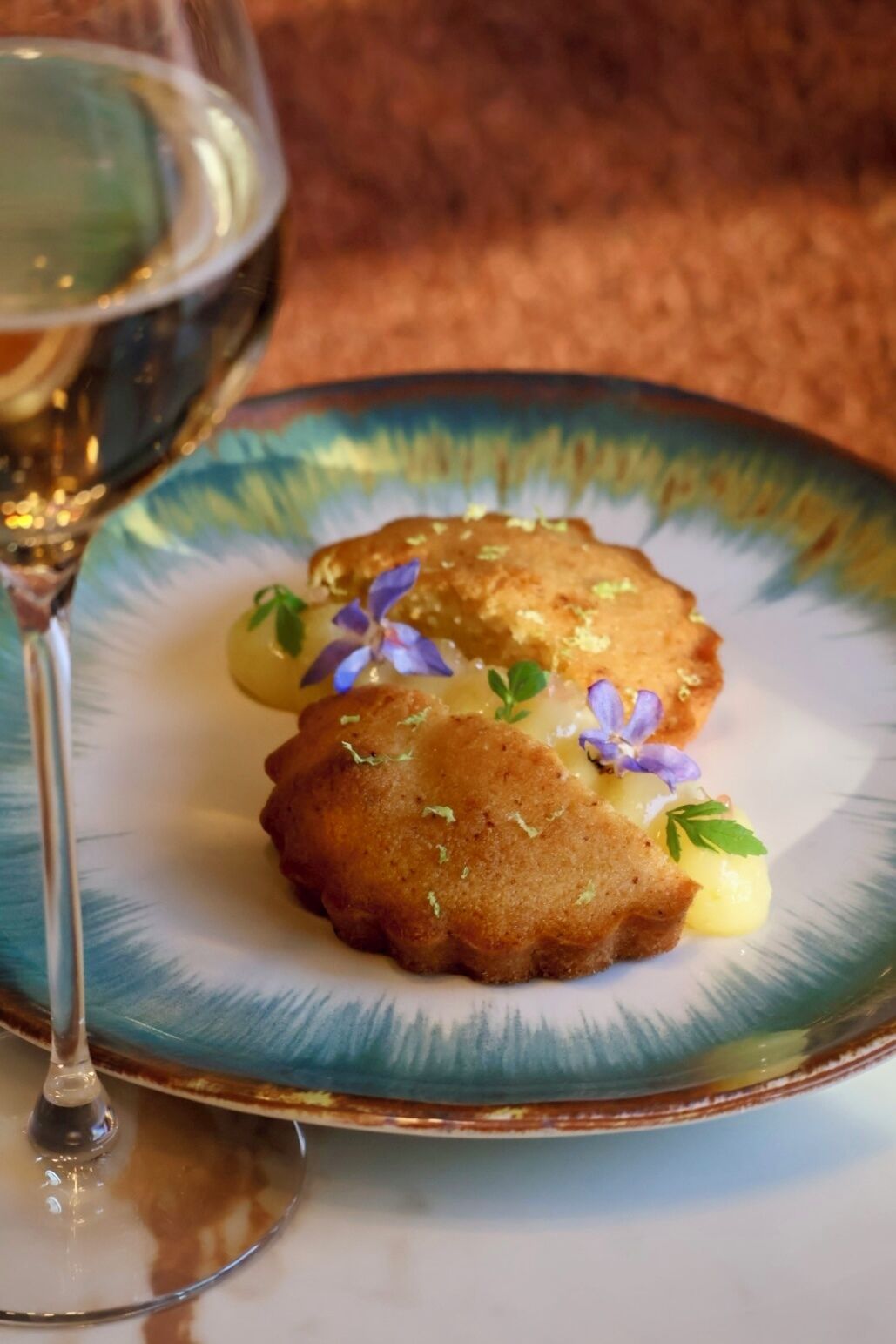 A plate with two golden-brown items, possibly fried pastries, garnished with small purple flowers and green leaves, accompanied by a glass of white wine.