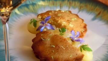 A plate with two golden-brown items, possibly fried pastries, garnished with small purple flowers and green leaves, accompanied by a glass of white wine.