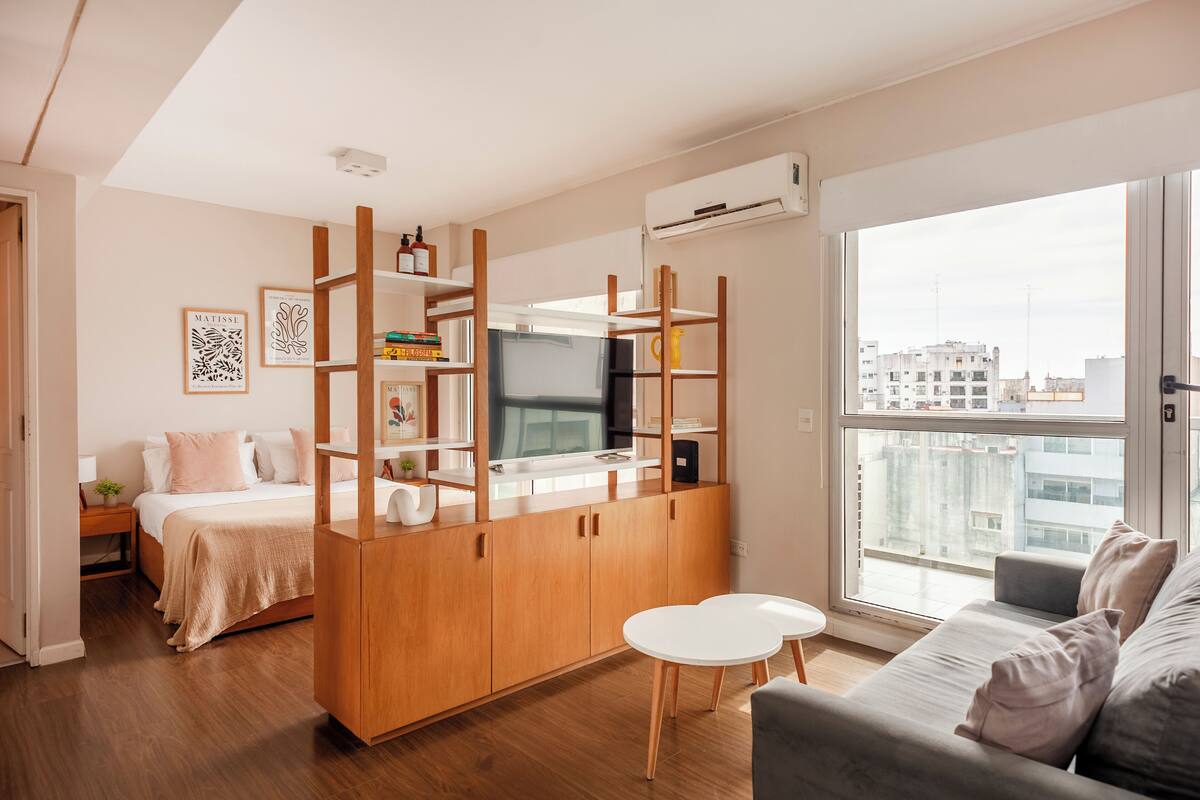 A modern apartment with a kitchenette, a bed, a sofa, and a large window offering a city view.