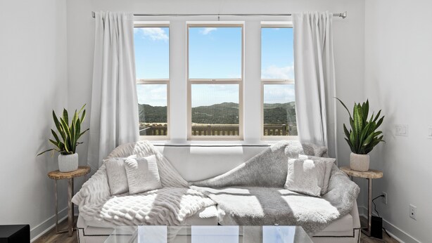 Una sala de estar moderna con un ventanal grande, un sofá blanco, dos mesitas de noche con plantas y vistas a colinas.