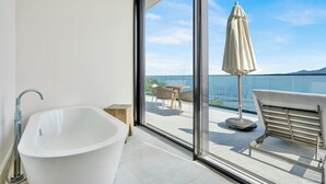 Une salle de bain moderne avec une baignoire sur pieds, un tabouret en bois et une vue sur un balcon agrémenté d’une table, de chaises et d’un parasol.