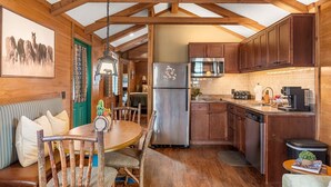 A rustic cabin interior with a wooden dining table, chairs, a kitchen with wooden cabinets, a refrigerator, a microwave, and a hanging light fixture.