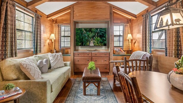 A cozy cabin interior with a wooden ceiling, a large window with curtains, a sofa, a dining table with chairs, and a small side table.
