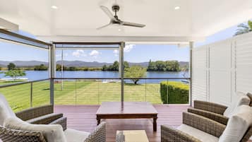 A covered outdoor deck with wicker furniture, a table, and a view of a lake and mountains.