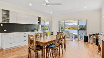 A modern kitchen with white cabinets, a wooden dining table, and chairs. There is a view of a lake and mountains through large sliding glass doors.