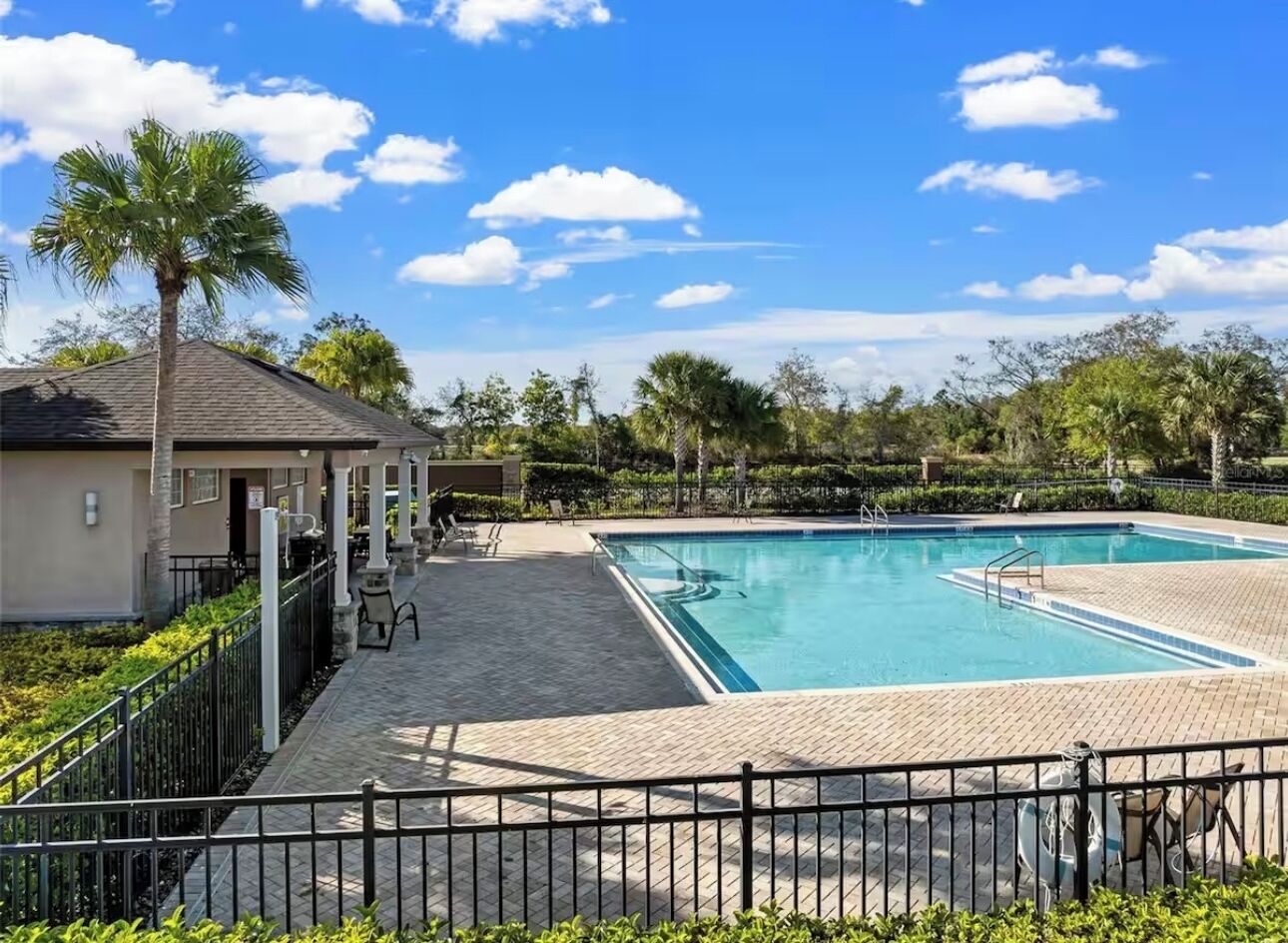 Una piscina con un borde de baldosas, rodeada por una valla y palmeras al fondo.
