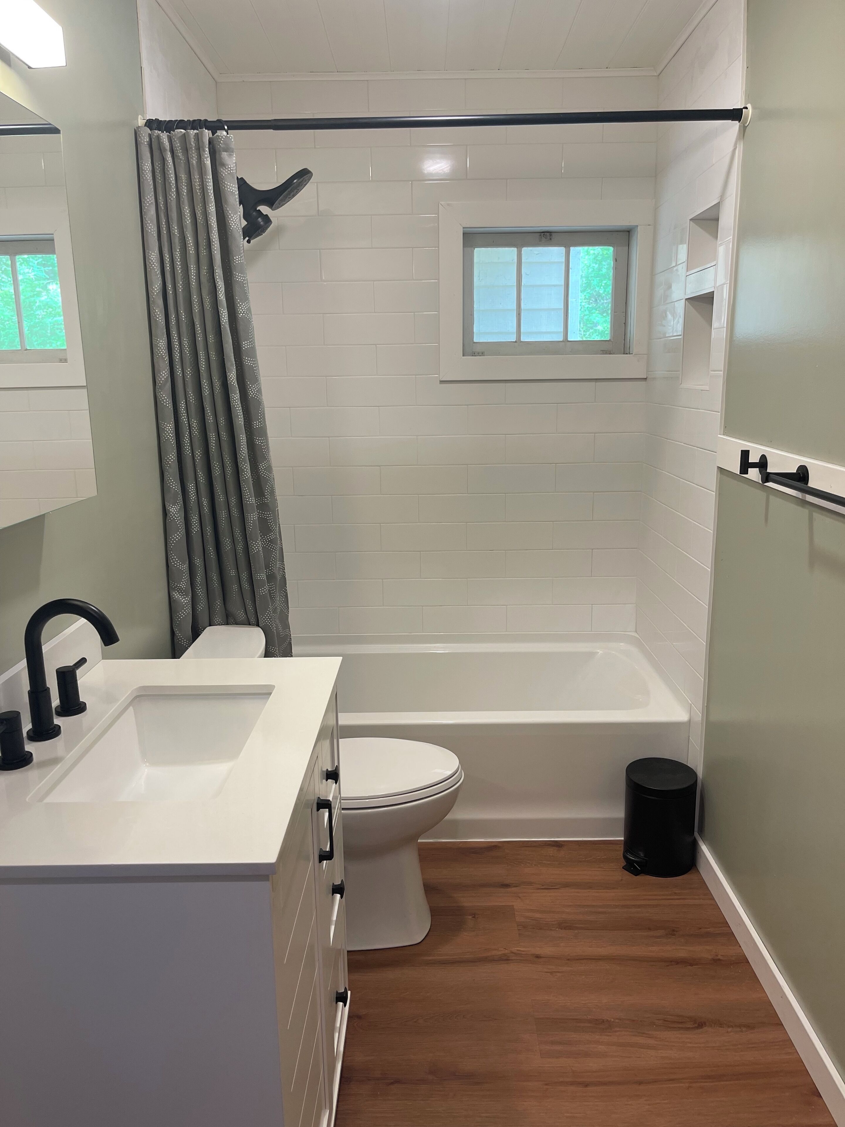Une salle de bain avec un lavabo, des toilettes et une baignoire, tous de couleur blanche. Il y a une fenêtre au-dessus de la baignoire et un rideau de douche.