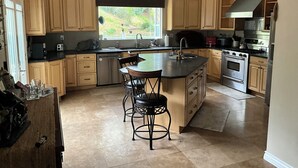 A modern kitchen with wooden cabinets, a central island with bar stools, stainless steel appliances, and a window providing natural light. - Oceanside tranquility (Oceanside)