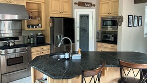 A modern kitchen with wooden cabinets, stainless steel appliances, a large island with bar stools, and a pantry door. - Oceanside tranquility (Oceanside)