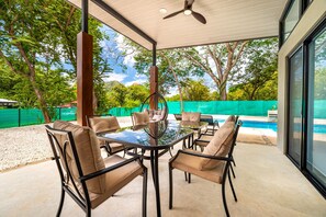 A covered outdoor dining area with a glass table and chairs, a ceiling fan, and a view of a swimming pool and trees.