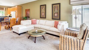 Ein Wohnzimmer mit einem Ecksofa, einem Glastisch, Korbstühlen, einem Essbereich mit Holzsesseln und gerahmten Pflanzenbildern an der Wand.