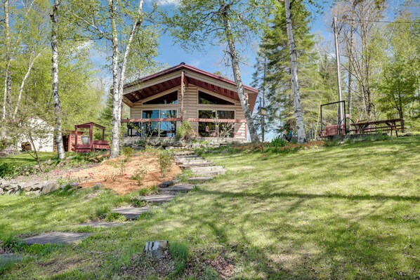Une cabane dotée d’une grande terrasse et d’un porche abrité, entourée d’arbres et bordée d’un sentier en pierre.
