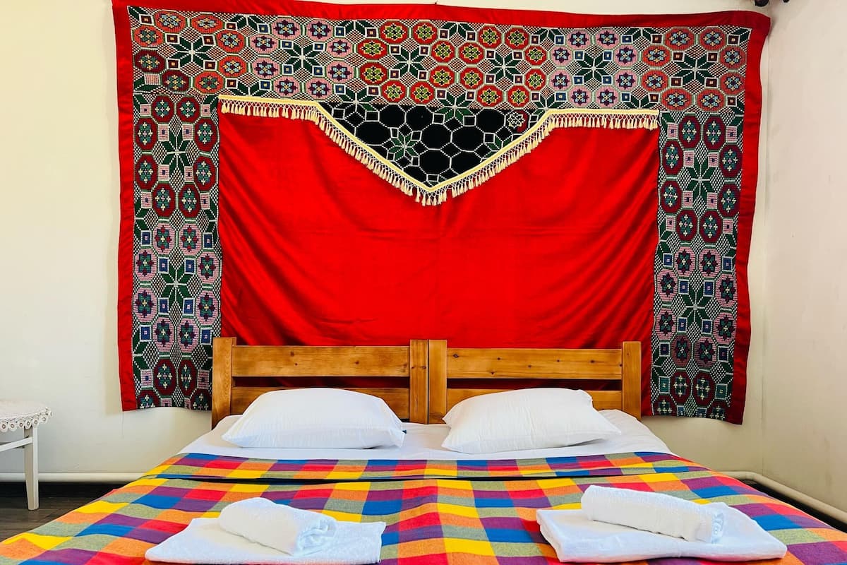 A bed with a colorful checkered bedspread, two white pillows, and a red and green patterned tapestry above.