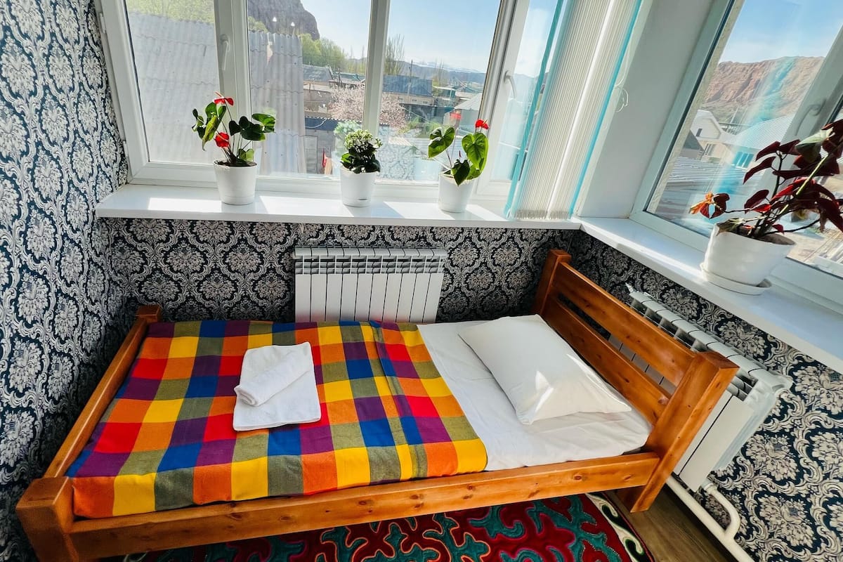 A small, neatly made bed with a colorful checkered bedspread and a white pillow. There are three potted plants on the windowsill, and the room has patterned wallpaper.