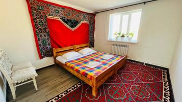 A bedroom with a wooden bed, a red patterned tapestry, a white chair, and a window with plants.