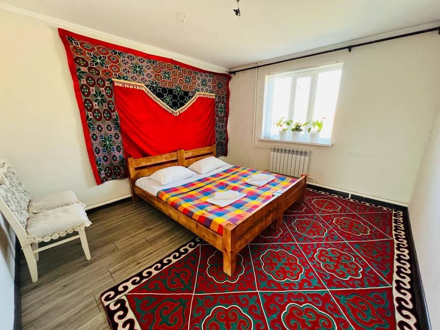 A bedroom with a wooden bed, a red patterned tapestry, a white chair, and a window with plants.