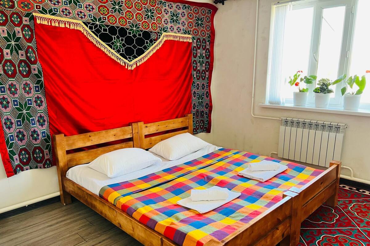 A wooden bed with a colorful checkered bedspread, white pillows, and a red curtain with a geometric pattern.