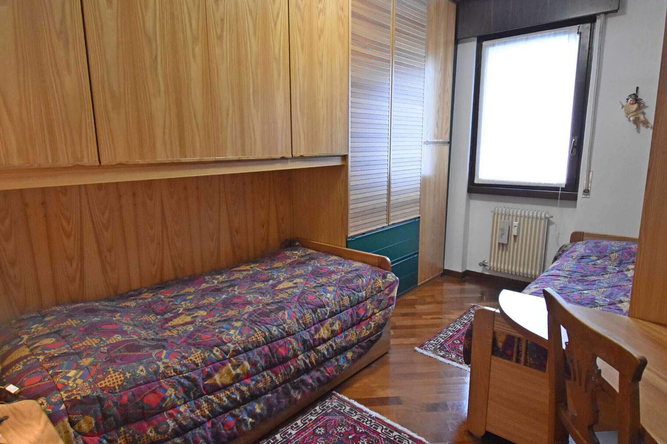 A small, compact room with a single bed, a wooden wardrobe, a window with blinds, and a radiator.