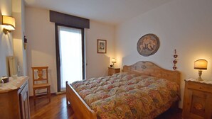 A bedroom with a wooden bed, a chair, a nightstand, and a wall-mounted lamp. - Presanella Alpine Apartment Mhd (Madonna di Campiglio)