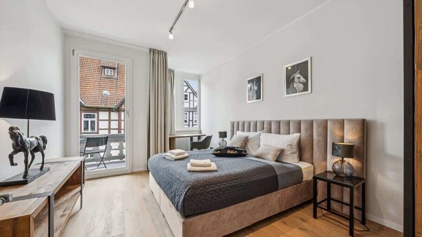 Ein modernes Schlafzimmer mit einem großen Bett, einem Schreibtisch und Blick auf ein Gebäude durch ein Fenster.