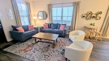 A living room with a sofa set, a coffee table, a white armchair, a small round table with chairs, and a decorative wall piece.