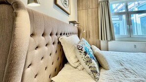 Un dormitorio con cabecera capitonada, cama con almohadones estampados, lámpara de noche, cuadro enmarcado en la pared y ventana con cortinas.