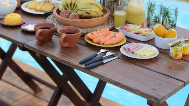 A wooden table with a variety of breakfast items including fruits, pastries, and beverages, set outdoors near a pool. - Vila Orun - Guest house (Barreirinhas)