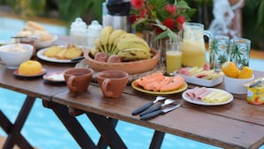 A breakfast table with various food items and drinks, set outdoors with a pool and palm trees in the background. - Vila Orun - Guest house (Barreirinhas)