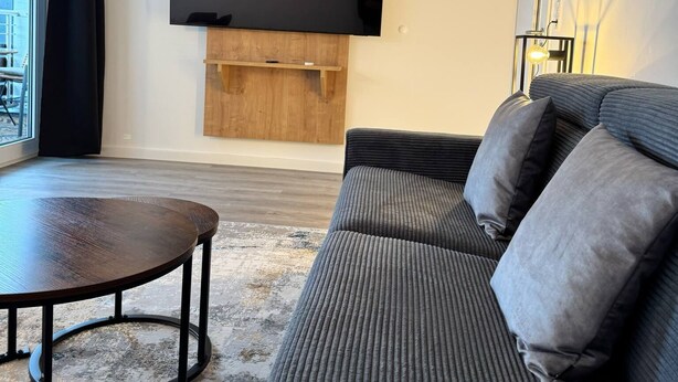 Een moderne woonkamer met een grote flatscreen televisie bevestigd aan een houten paneel, een donkergrijze bank met kussens, een ronde salontafel en uitzicht op het groen door een groot raam met zwarte gordijnen.