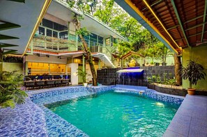 A swimming pool with a waterfall feature, surrounded by a covered outdoor area with seating and a staircase leading to an upper level. - Clarence Retreat Goa (Marra)