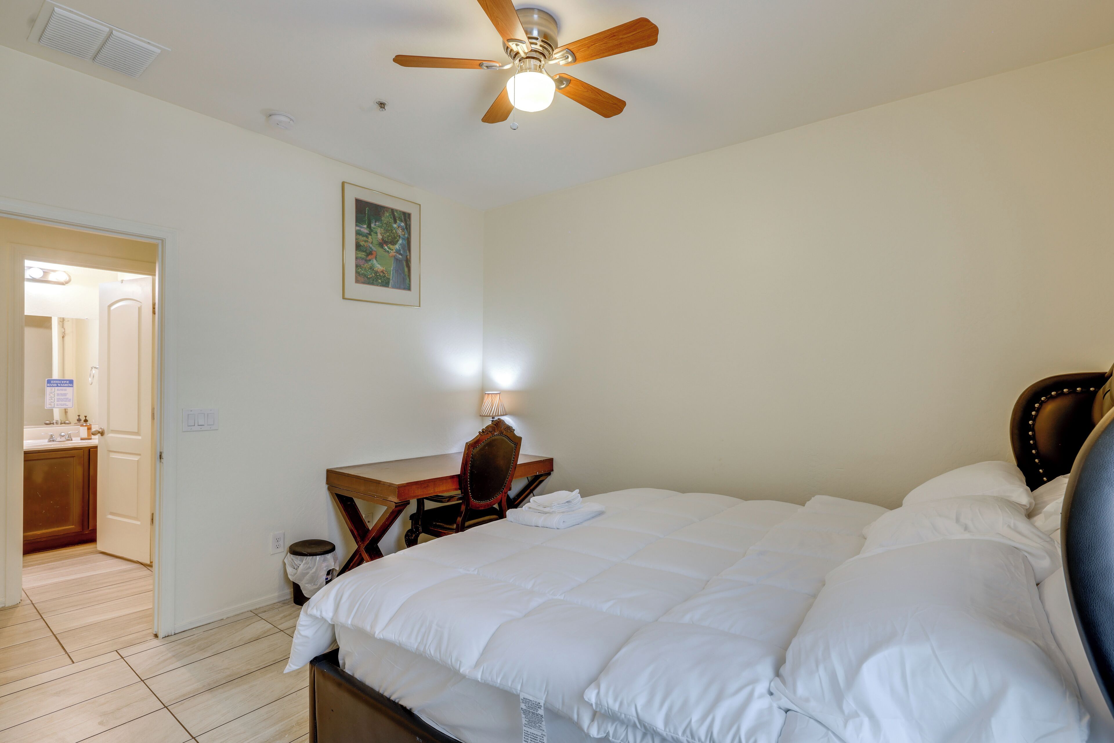 Een slaapkamer met een bed, een bureau, een stoel, een plafondventilator en een deur naar een badkamer.