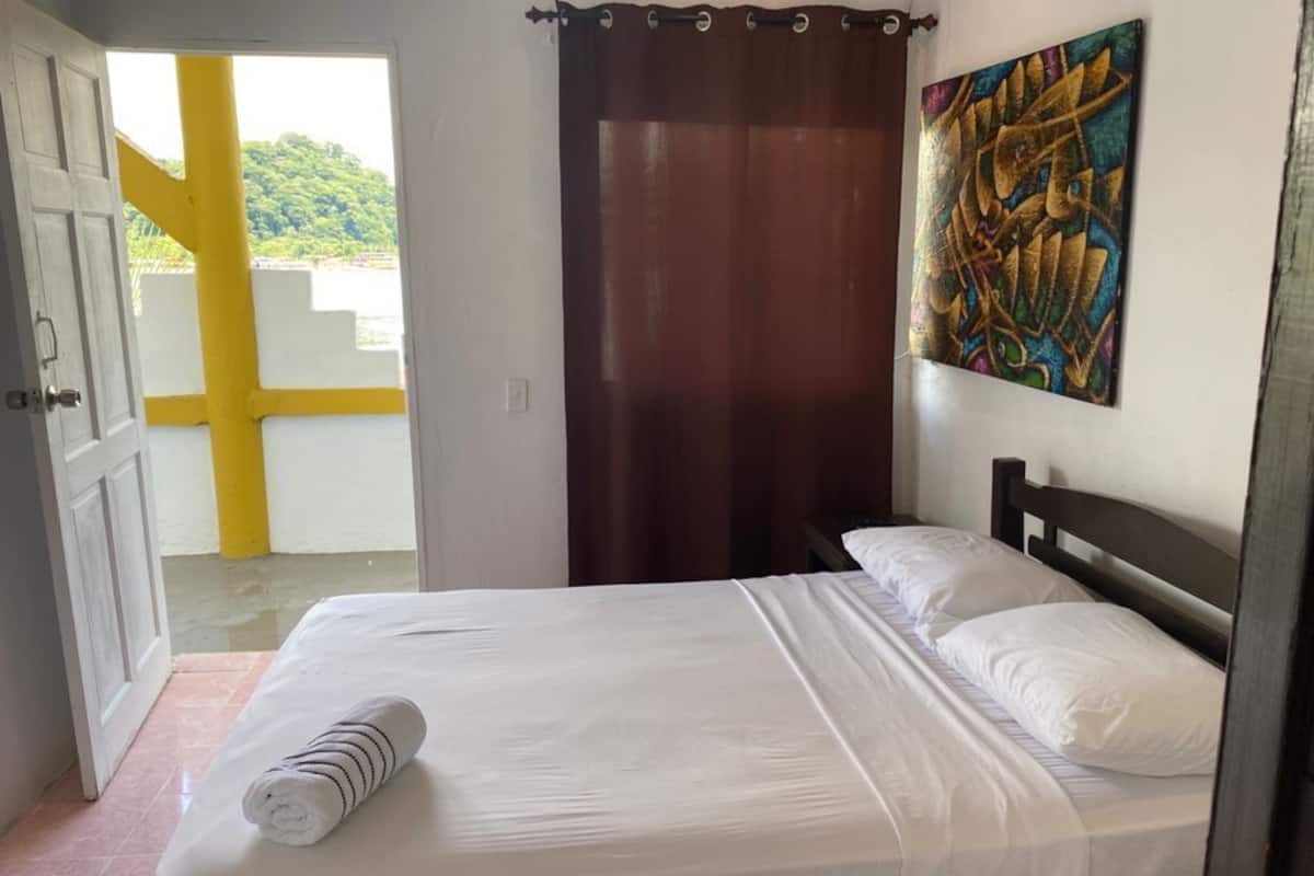 A bedroom with a bed, a painting on the wall, and a view of a yellow structure outside.