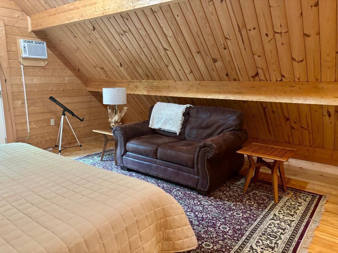 Una habitación acogedora con techo de madera, un sofá de cuero marrón, una mesita, una lámpara y una cama con colcha acolchada.