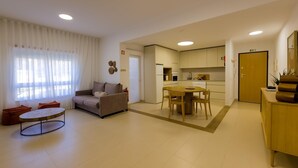 A modern living room with a sofa, a round coffee table, and a dining table with chairs. There is a kitchen area with cabinets and a refrigerator.