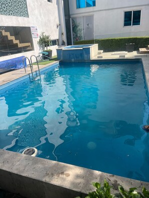A swimming pool with clear blue water, surrounded by a concrete deck and a building with a white facade.