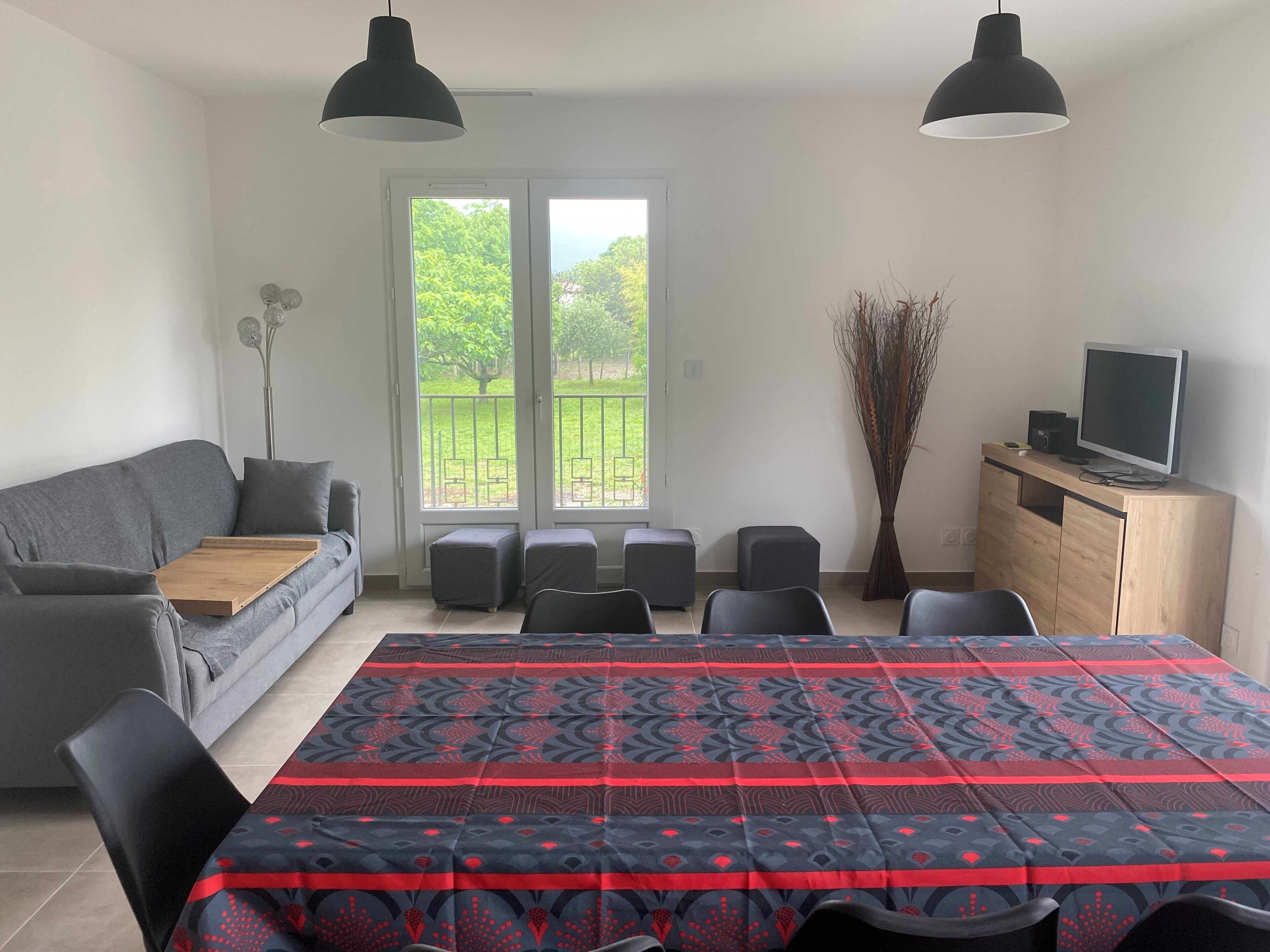Un salon moderne avec un canapé gris, une table basse en bois, un tapis à motifs, un téléviseur posé sur un meuble en bois, et une vue sur la verdure à travers la porte vitrée.