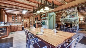A rustic cabin interior with a wooden dining table, chairs, a kitchen area, and a stone fireplace. - The Alibi Cabin - Pool, Hot Tub & Private Trails - Secluded 17-Acre Retreat (Kensington)