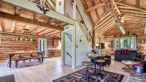 A log cabin interior with a dining area, kitchen, and living space, featuring wooden beams, a staircase, and a view of the outdoors. - The Alibi Cabin - Pool, Hot Tub & Private Trails - Secluded 17-Acre Retreat (Kensington)