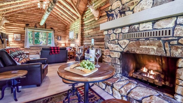 A cozy log cabin interior with a stone fireplace, wooden furniture, and a dining table set with food and wine. - The Alibi Cabin - Pool, Hot Tub & Private Trails - Secluded 17-Acre Retreat (Kensington)