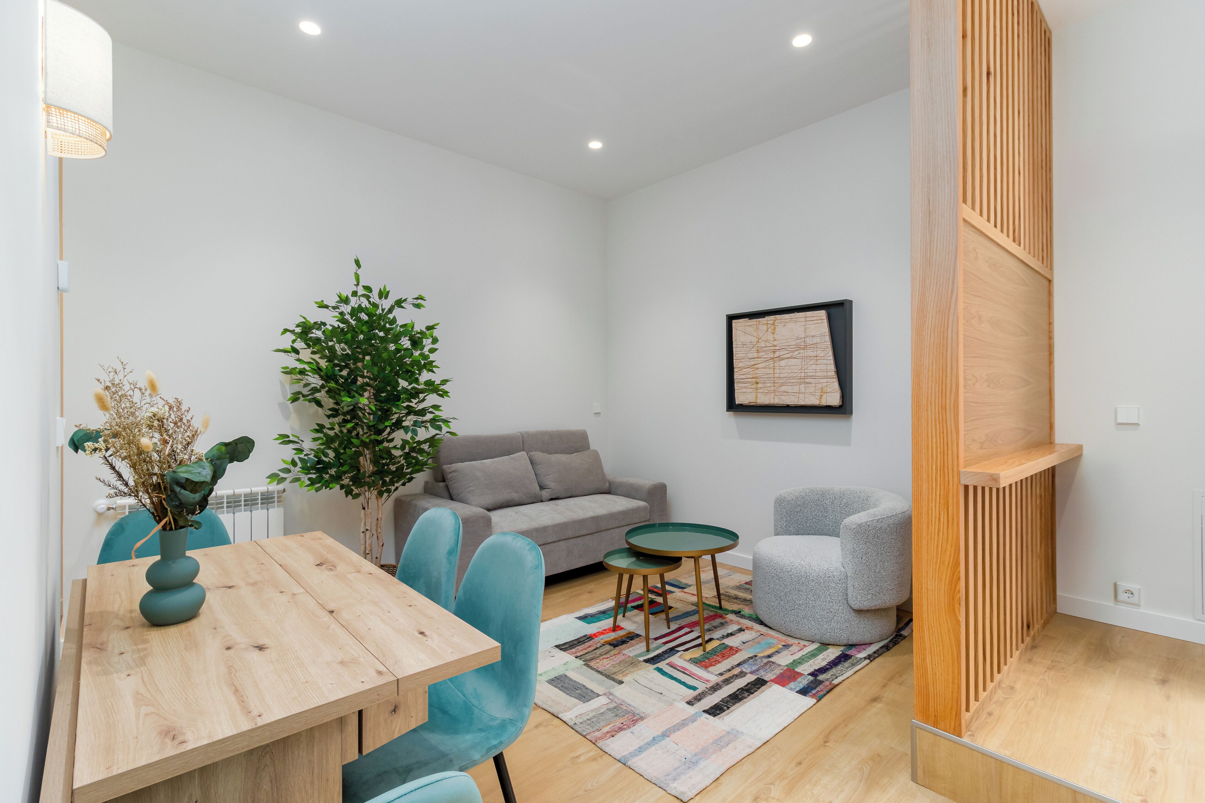 Een moderne woonkamer met een houten eettafel, teal stoelen, een grijze bank, een kleine ronde tafel en een kamerplant.
