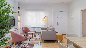 Un salon moderne avec un canapé, des fauteuils, une table basse et un téléviseur. On y trouve également une plante, une lampe et un radiateur.