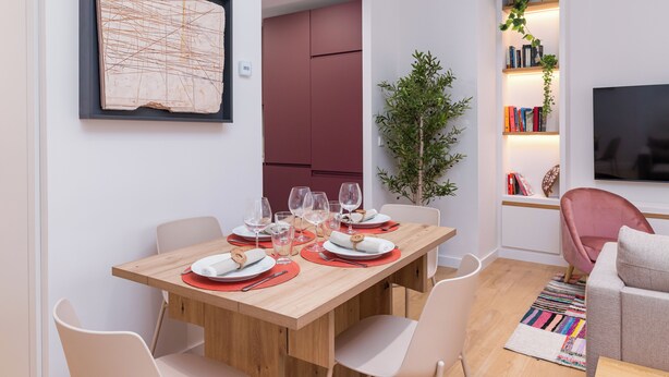 Un coin repas avec une table en bois dressée pour quatre personnes, entourée de chaises modernes. On aperçoit une bibliothèque avec des livres et une plante au fond.