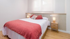 Un lit bien fait, recouvert d’une couverture rouge, une table de chevet avec une lampe et un mur orné d’un motif.